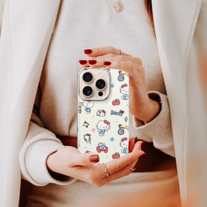 May include: A phone case with a repeating pattern of Hello Kitty characters, apples, stars, and other cute illustrations on a cream background. The case has a clear border and is held in front of a person.