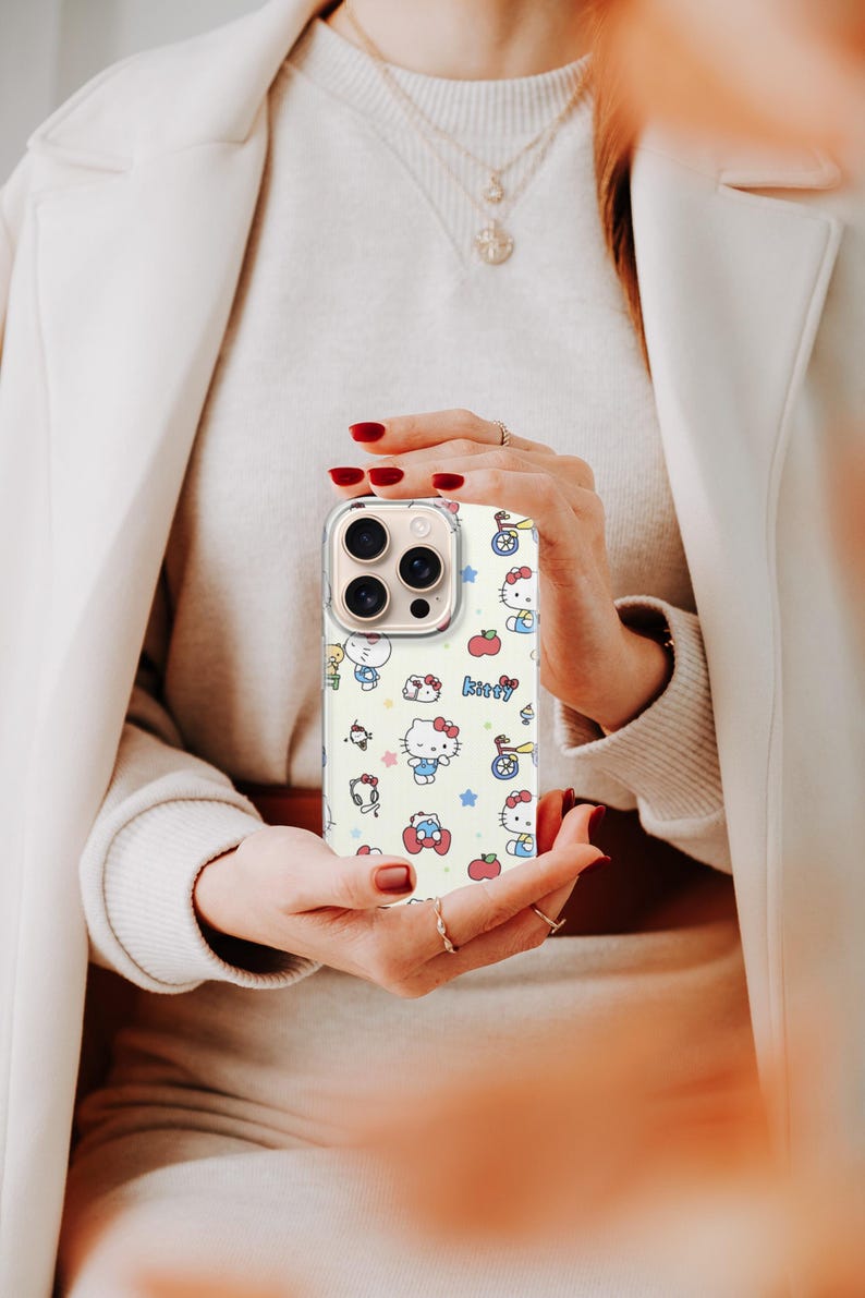 May include: A phone case with a repeating pattern of Hello Kitty characters, apples, stars, and other cute illustrations on a cream background. The case has a clear border and is held in front of a person.