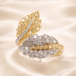May include: A leaf-shaped ring with a gold band and two diamond-encrusted leaf designs. One leaf is yellow, the other is white, and both are adorned with numerous small diamonds. The ring is set against a soft, off-white background.