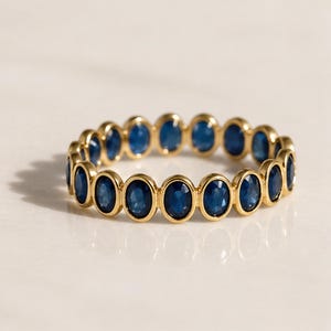 May include: A gold ring with a continuous band of oval, deep blue gemstones. The gemstones are set in individual bezels. The ring is presented on a white surface, showcasing its design and colour.