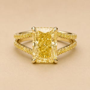 May include: A yellow diamond ring with a split shank design. The ring features a large, rectangular yellow diamond as the centerpiece, with smaller yellow diamonds set along the band. The ring is set in a gold band.