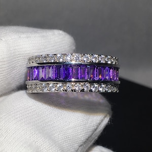 May include: A silver ring with a band of rectangular, deep purple gemstones. The ring is bordered by two rows of small, round, clear gemstones. The ring is shown on a white surface.