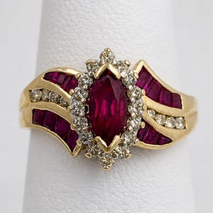 May include: A gold ring featuring a marquise-cut ruby center stone surrounded by diamonds. The ring has additional ruby accents and diamond clusters on the band, set in a gold band. A classic and elegant jewelry piece.