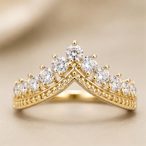 Pode incluir: Um anel de ouro com uma banda em forma de V, textura granulada e uma fila de pedras redondas, claras e facetadas ao longo da borda superior. As pedras estão dispostas em forma de coroa, com a pedra central no ponto mais alto.
