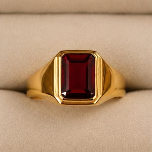 May include: A gold-colored ring with a rectangular, deep red gemstone. The ring has a classic design with a thick band and a bezel setting for the gemstone. The gemstone is a rich, dark red color.