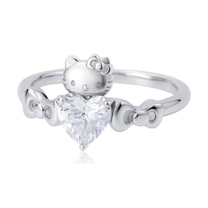 May include: A silver ring featuring a heart-shaped clear gemstone. The ring has a Hello Kitty head on top and bow accents on the sides. The ring is a piece of jewelry.