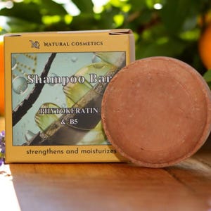 Peut inclure: Un pain de shampooing rond et brun à côté d'une boîte de shampooing Natural Cosmetics. La boîte est beige et porte les mots "Shampoo Bar" et "PhytoKeratin & B5". Des oranges et des fleurs violettes sont en arrière-plan.