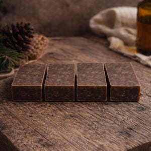 May include: Four rectangular brown bars of soap are arranged on a rustic wooden surface. The soap has a textured appearance. The background includes a pine cone, a bottle, and a cloth, suggesting a natural or spa theme.