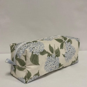 Puede incluir: Estuche rectangular con un estampado floral de flores de hortensia azul claro y hojas verdes sobre un fondo crema. Tiene una cremallera azul claro y un pequeño lazo de tela. Aproximadamente 20 cm de largo.