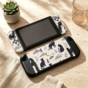 May include: A handheld gaming console featuring a custom skin with a pattern of black cats, crescent moons, stars, lit candles, and green leaves on a cream background. The console includes a matching protective case with the same cat-themed design.