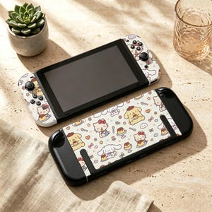 May include: A handheld gaming console featuring a white skin with a repeating pattern of Sanrio characters including Hello Kitty, Pompompurin, and Cinnamoroll alongside small pudding desserts, hearts, and flowers on the controllers and back panel.
