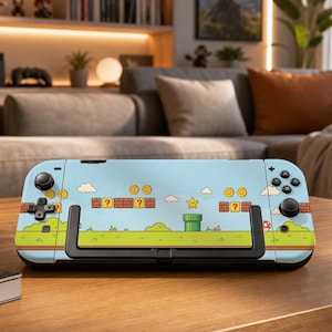 May include: A handheld gaming console featuring a light blue skin with a graphic print of a video game landscape. The design includes yellow coins, brown question mark blocks, a green pipe, a yellow star, and small green bushes on a grassy field.