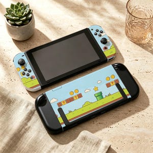 May include: A handheld gaming console with a light blue skin featuring a video game landscape with green grass, brown brick blocks, gold coins, a green pipe, and a yellow star. The console has a black screen and black buttons on the controllers.