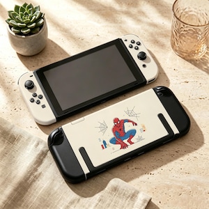 May include: A handheld gaming console with a white and black frame, featuring a screen and control buttons. Beside it lies a matching console case in cream, black, and red, decorated with a graphic illustration of Spider-Man and spiderwebs.