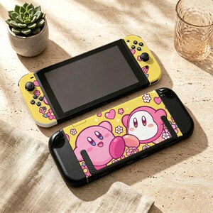 May include: A handheld video game console featuring a yellow skin with pink flowers and hearts. A matching protective case displays a graphic of two pink Kirby characters surrounded by pink hearts and white flowers on a bright yellow background.