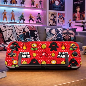 Puede incluir: Una funda roja para consola de juegos portátil con un patrón de puntos blancos, el texto Super Mario y varios iconos que incluyen flores de fuego, monedas de oro, gorras de Mario, llamas y siluetas negras de personajes Bowser en el frente.