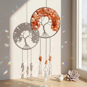 Puede incluir: Un adorno de pared artesanal con un diseño de árbol de alambre adornado con piedras preciosas naranjas. El árbol está suspendido dentro de un aro plateado, con cadenas colgantes de cuentas y prismas de cristal. La luz del sol proyecta una sombra en forma de árbol.