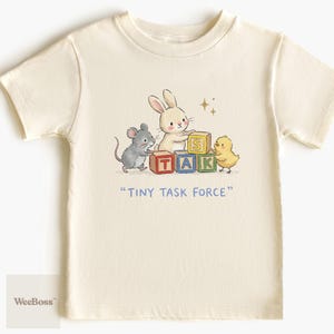 May include: Cream-colored youth t-shirt featuring a whimsical illustration of a bunny, mouse, and chick arranging TAK blocks. The text "TINY TASK FORCE" is below the image. The shirt is made of cotton.