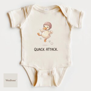 May include: Cream-colored baby onesie with a cartoon duck wearing a helmet and the text "QUACK ATTACK." The duck is depicted mid-stride, with one foot raised. The WeeBoss logo is in the lower left corner.