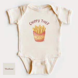May include: Cream-colored baby onesie with the text "CHIPPY THIEF" above a cartoon illustration of a smiling container of french fries. The fries are golden yellow, and the container is pink and red.