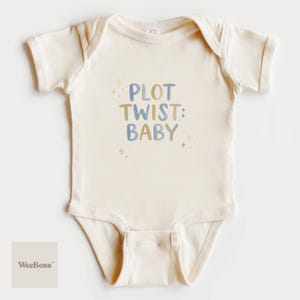 May include: Cream-colored baby onesie with the text "PLOT TWIST: BABY" in blue and tan lettering. The onesie has short sleeves and a snap closure. Small star graphics are scattered around the text. The WeeBoss logo is in the bottom left corner.