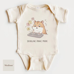 May include: Cream-colored baby onesie featuring a cartoon hamster with a worried expression, typing on a keyboard. The text "DEADLINE PANIC MODE" is below the hamster. The design includes exclamation points and warning symbols.