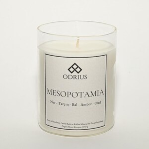 May include: A clear glass candle holder with a white candle inside. The candle has a label with the word "MESOPOTAMIA" and the brand name "ODRIUS". The label also lists ingredients and the candle weighs 150g.