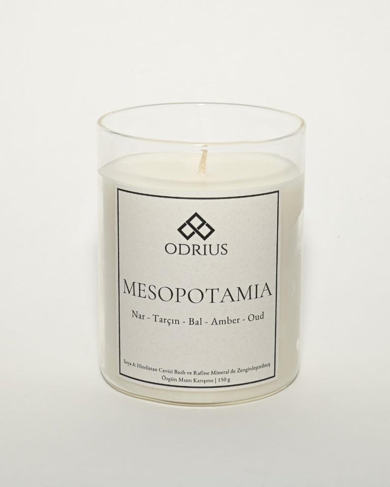 May include: A clear glass candle holder with a white candle inside. The candle has a label with the word "MESOPOTAMIA" and the brand name "ODRIUS". The label also lists ingredients and the candle weighs 150g.