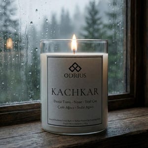 Op de afbeelding: Een brandende kaars in een heldere glazen pot met een wit etiket. Het etiket toont "ODRIUS KACHKAR" met tekst in een vreemde taal. De kaars staat op een houten vensterbank, met een regenachtige bosscène door het raam.