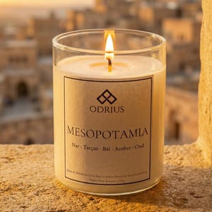 May include: A lit candle in a clear glass jar. The candle has a label with the word "MESOPOTAMIA" and the brand name "ODRIUS". The candle is on a stone surface with a blurred background of buildings.
