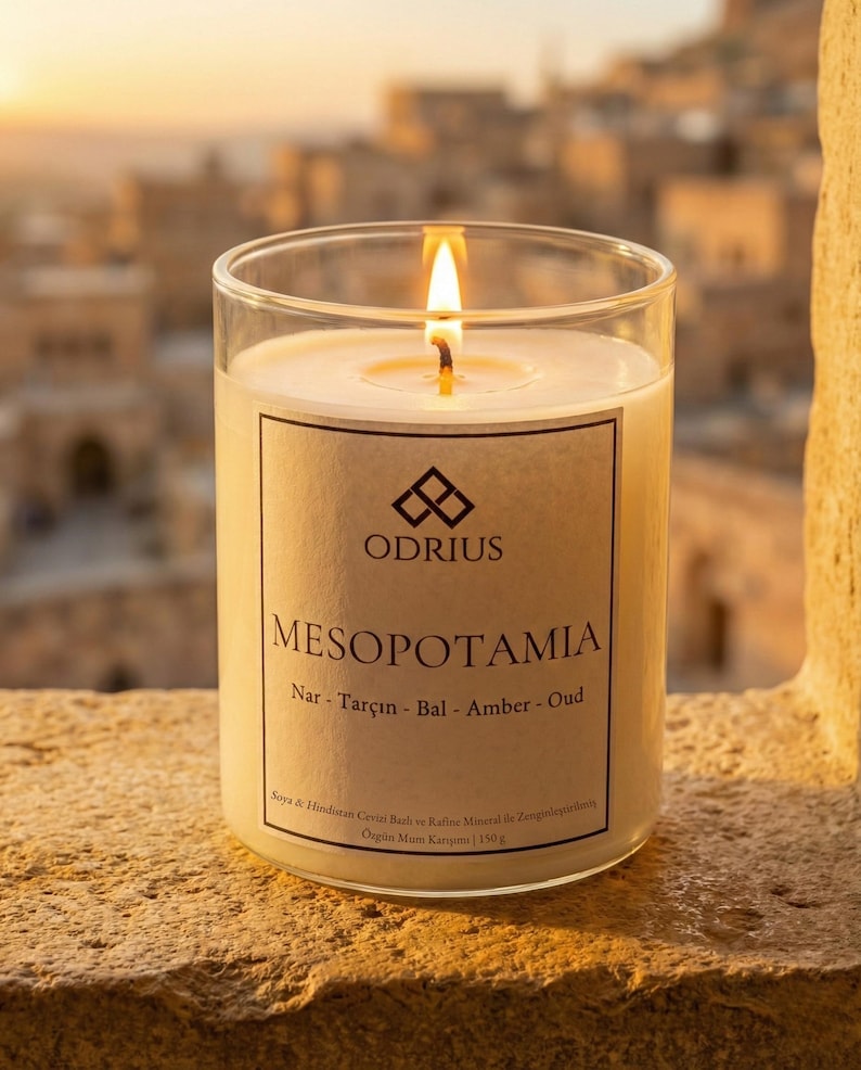 May include: A lit candle in a clear glass jar. The candle has a label with the word "MESOPOTAMIA" and the brand name "ODRIUS". The candle is on a stone surface with a blurred background of buildings.