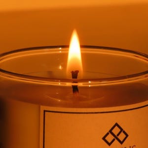 May include: A lit candle in a clear glass container. The flame is bright yellow and orange, with a black wick. The candle has a white label with the word "ODRIUS" and a geometric logo.
