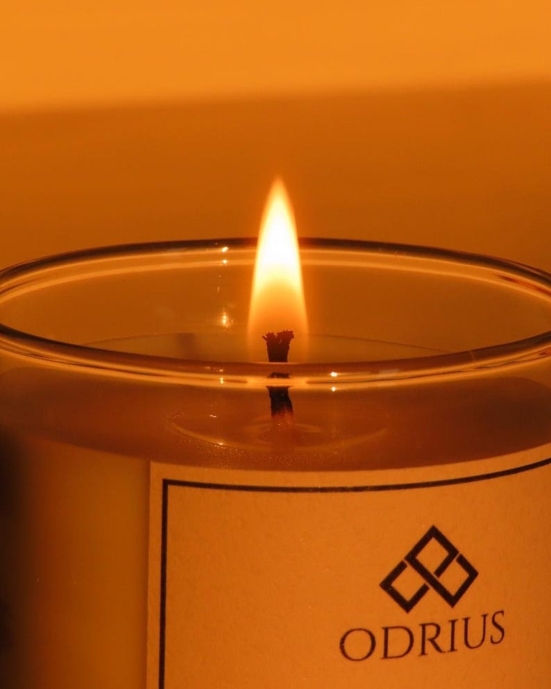 May include: A lit candle in a clear glass container. The flame is bright yellow and orange, with a black wick. The candle has a white label with the word "ODRIUS" and a geometric logo.