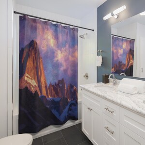 May include: A shower curtain featuring a mountain landscape with a sunset sky. The mountains are shades of brown and orange, with a purple and yellow sky. The curtain is hanging in a bathroom with white cabinets and a marble countertop.