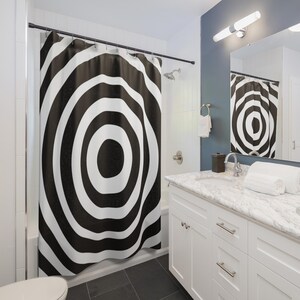 Puede incluir: Cortina de ducha con un patrón de círculos concéntricos en blanco y negro. La cortina cuelga en un baño con gabinetes blancos, una encimera de mármol y un espejo que refleja el diseño. El baño tiene un diseño moderno.