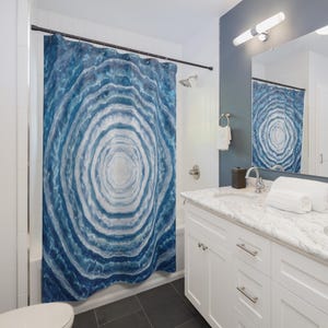 May include: A blue and white shower curtain with a concentric circle design, resembling a cross-section of a tree trunk. The curtain hangs in a bathroom with white cabinetry, a marble countertop, and a dark gray tiled floor. A matching design is reflected in the mirror.