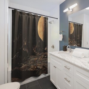 May include: A shower curtain depicting a celestial scene with a large, textured, golden-orange planet and a dark, rocky terrain. The curtain is in a bathroom with white cabinets and a marble countertop.