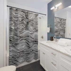 Puede incluir: Una cortina de ducha en blanco y negro con un patrón ondulado a rayas. La cortina cuelga en un baño con un tocador blanco, un espejo y un cabezal de ducha. El patrón se asemeja a rayas de cebra.