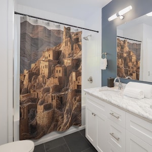 May include: A shower curtain featuring a detailed print of a mountain village. The image showcases a cluster of buildings nestled into a rocky hillside, with warm, earthy tones dominating the scene. The curtain is hung in a bathroom setting with white fixtures and a dark floor.