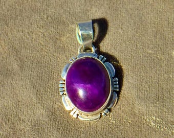 May include: A silver pendant with an oval, deep purple gemstone. The gemstone is set in a decorative silver frame with a bail at the top for a chain. The pendant is displayed against a neutral background.