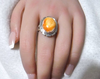 May include: A silver ring featuring a large, oval-shaped spiny oyster shell cabochon. The shell displays vibrant orange and yellow hues with a textured surface. The ring has a decorative, scalloped bezel and a simple band.