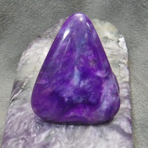 May include: A polished, triangular-shaped purple stone with white and blue veining. The stone sits on a larger, rectangular piece of stone with a similar color pattern. The overall effect is a striking display of natural beauty.