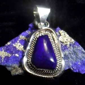 Puede incluir: Un colgante de plata con una piedra preciosa de color púrpura oscuro engastada en un engaste de plata. El colgante está rodeado por un diseño de cuerda de plata.