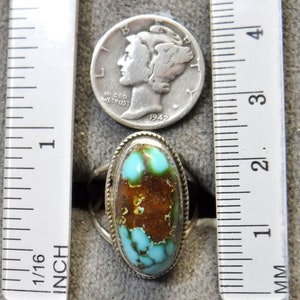K&ouml;nnte beinhalten: Ein silberner Ring mit einem t&uuml;rkisfarbenen Stein, der in einer ovalen Form gefasst ist. Der Stein ist eine Mischung aus blauen und braunen Farben. Der Ring liegt neben einem Lineal zur Gr&ouml;&szlig;enreferenz.