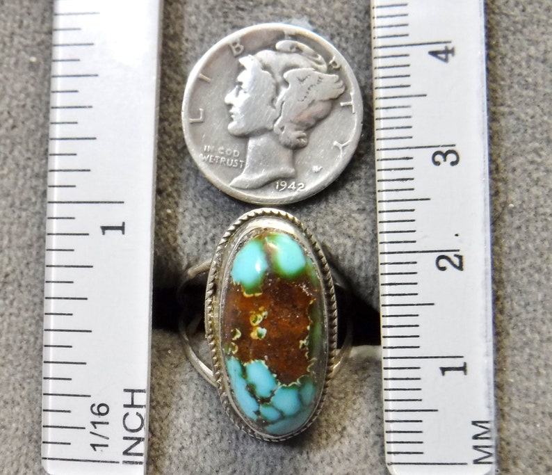 K&ouml;nnte beinhalten: Ein silberner Ring mit einem t&uuml;rkisfarbenen Stein, der in einer ovalen Form gefasst ist. Der Stein ist eine Mischung aus blauen und braunen Farben. Der Ring liegt neben einem Lineal zur Gr&ouml;&szlig;enreferenz.