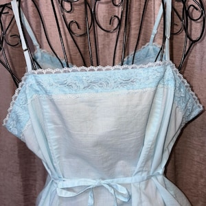 May include: A light blue slip dress with spaghetti straps and a tie waist. The dress features a delicate lace trim along the neckline and a soft, flowing fabric. The vintage-inspired design is displayed on a black metal dress form.