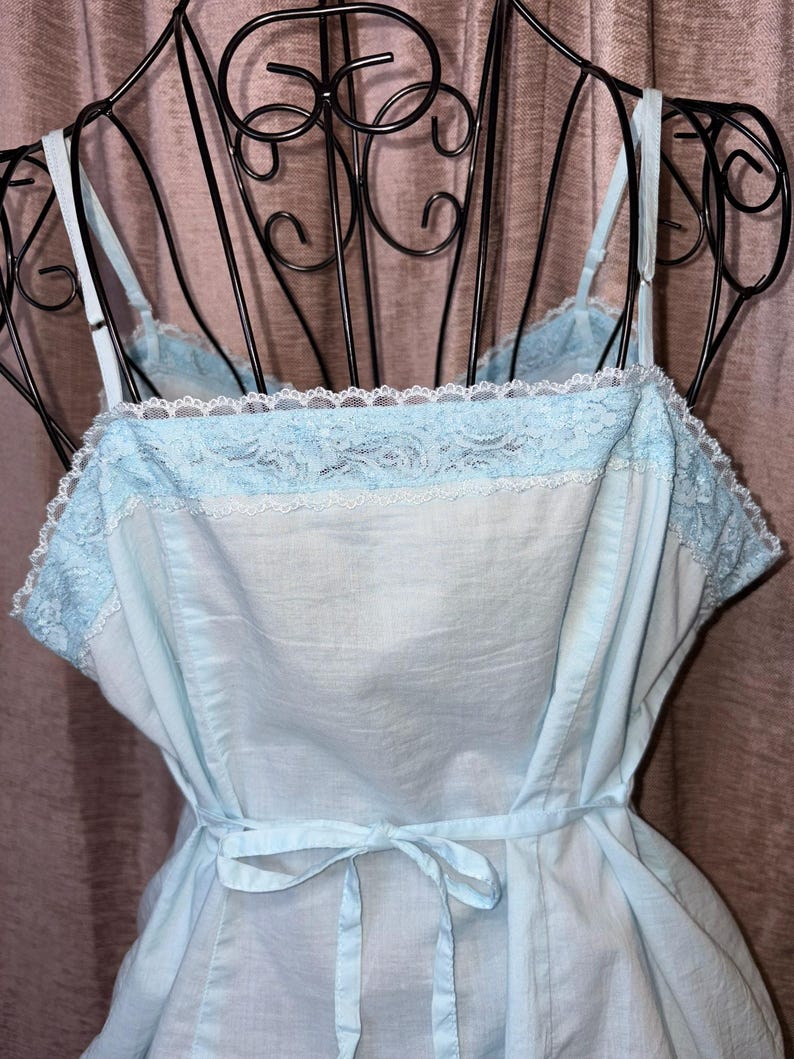 May include: A light blue slip dress with spaghetti straps and a tie waist. The dress features a delicate lace trim along the neckline and a soft, flowing fabric. The vintage-inspired design is displayed on a black metal dress form.
