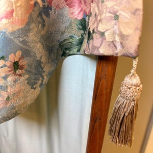 Puede incluir: Primer plano de una tela estampada con flores en tonos rosa, azul y verde. La tela est&aacute; drapeada sobre un poste de madera, con una borla beige colgando del borde. El dise&ntilde;o floral presenta grandes flores.