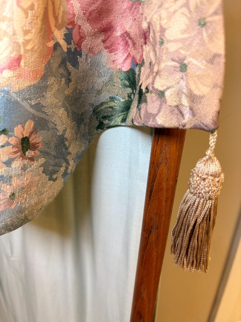 Puede incluir: Primer plano de una tela estampada con flores en tonos rosa, azul y verde. La tela est&aacute; drapeada sobre un poste de madera, con una borla beige colgando del borde. El dise&ntilde;o floral presenta grandes flores.