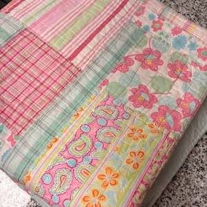 May include: A quilted fabric piece featuring a patchwork design with various patterns including pink and white plaid, light green stripes, floral motifs in pink and blue, and a paisley print in pink, yellow, and green, all stitched together.